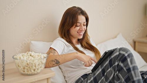 Young beautiful hispanic woman using touchpad eating popcorn sitting on bed at bedroom
