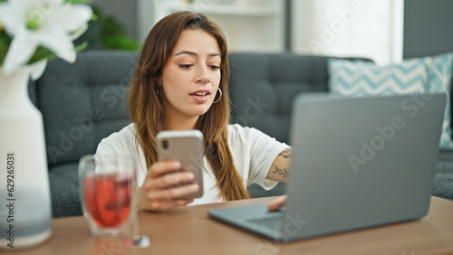 Young beautiful hispanic woman using laptop and smartphone sitting on floor at home