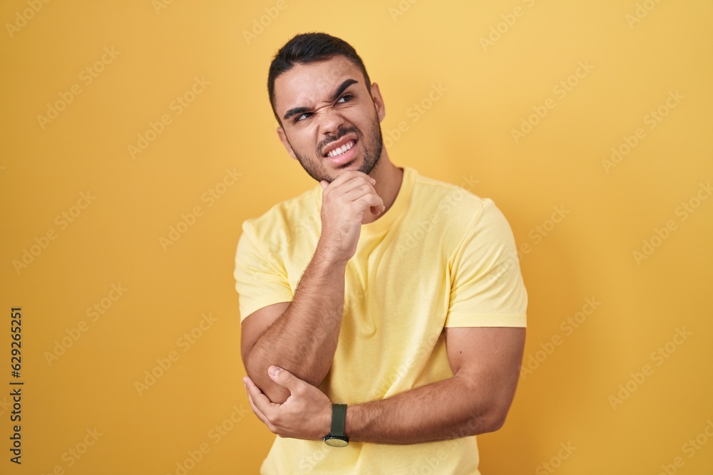 Young hispanic man standing over yellow background thinking worried about a question, concerned and nervous with hand on chin Young hispanic man standing over yellow background thinking worried about a question, concerned and nervous with hand on chin