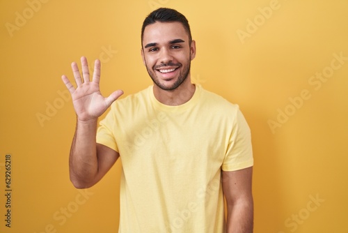 Young hispanic man standing over yellow background showing and pointing up with fingers number five while smiling confident and happy. © Krakenimages.com