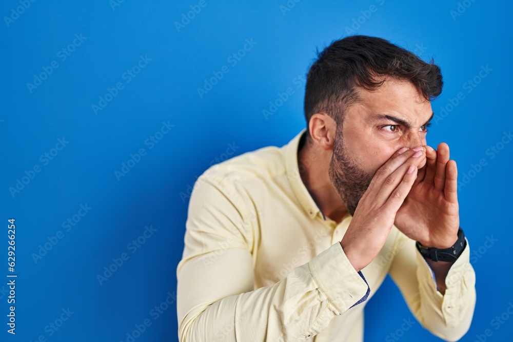 Handsome hispanic man standing over blue background shouting angry out loud with hands over mouth Handsome hispanic man standing over blue background shouting angry out loud with hands over mouth