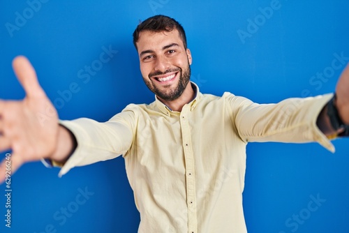 Handsome hispanic man standing over blue background looking at the camera smiling with open arms for hug. cheerful expression embracing happiness.