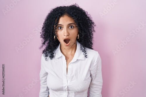 Hispanic woman with curly hair standing over pink background afraid and shocked with surprise and amazed expression, fear and excited face.