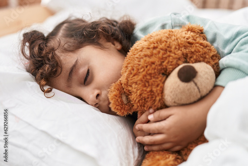 Adorable hispanic girl hugging teddy bear lying on bed sleeping at bedroom