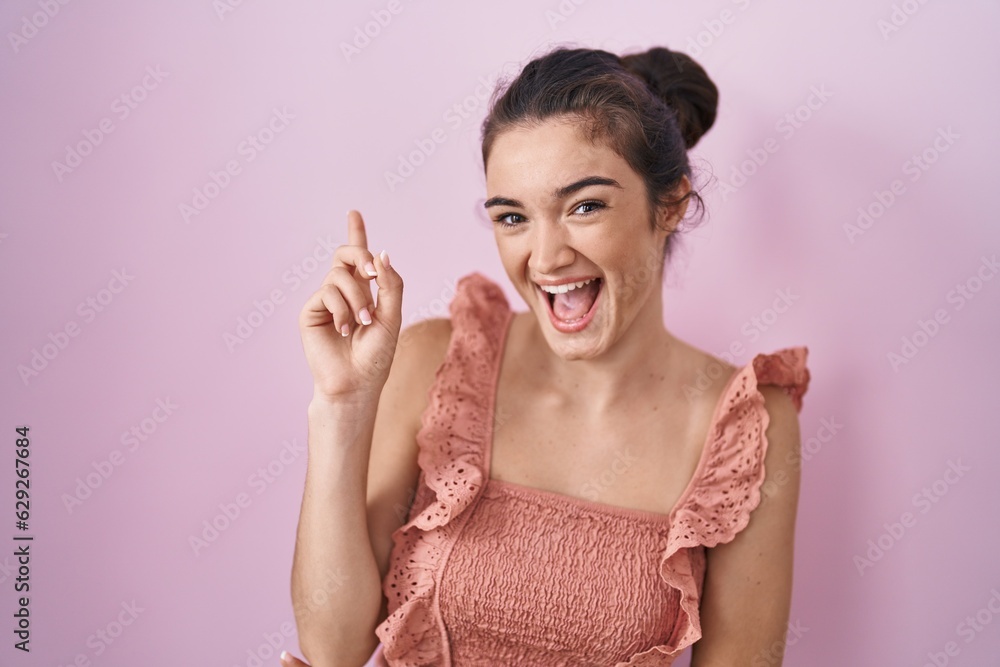Young teenager girl standing over pink background with a big smile on face, pointing with hand and finger to the side looking at the camera. Young teenager girl standing over pink background with a big smile on face, pointing with hand and finger to the side looking at the camera.