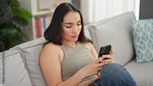 Young beautiful hispanic woman using smartphone sitting on sofa at home