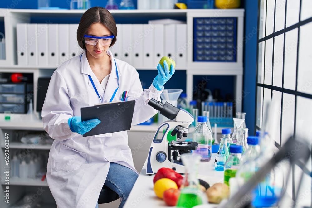 Young beautiful hispanic woman scientist holding apple reading document at laboratory Young beautiful hispanic woman scientist holding apple reading document at laboratory