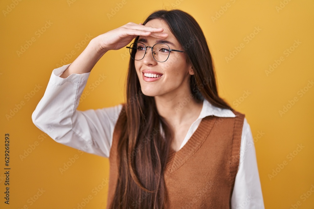 Young brunette woman standing over yellow background wearing glasses very happy and smiling looking far away with hand over head. searching concept. Young brunette woman standing over yellow background wearing glasses very happy and smiling looking far away with hand over head. searching concept.