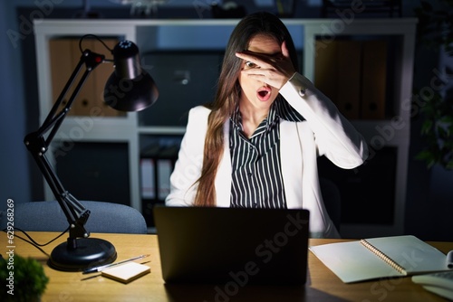Fotografía Young brunette woman working at the office at night with laptop peeking in shock covering face and eyes with hand, looking through fingers with embarrassed expression