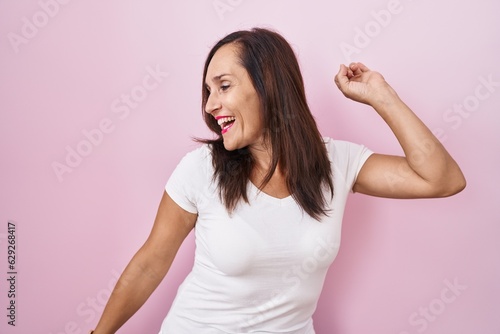 Middle age brunette woman standing over pink background dancing happy and cheerful  smiling moving casual and confident listening to music