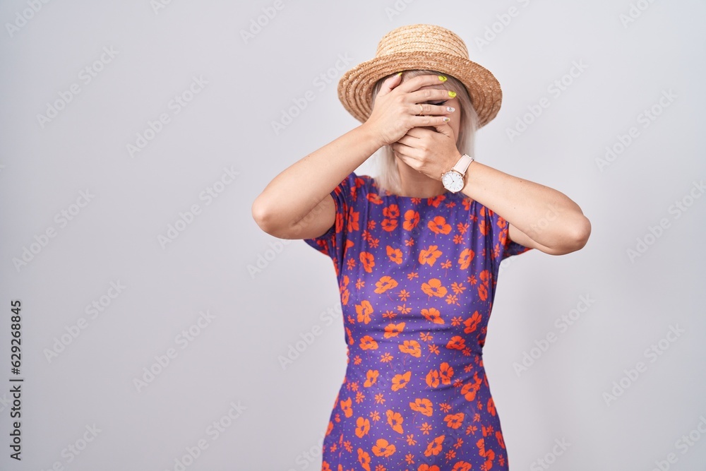 Young caucasian woman wearing flowers dress and summer hat covering eyes and mouth with hands, surprised and shocked. hiding emotion Young caucasian woman wearing flowers dress and summer hat covering eyes and mouth with hands, surprised and shocked. hiding emotion