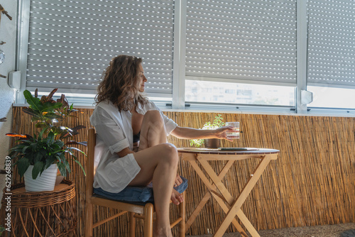 Obraz na płótnie Young caucasian woman in a white shirt sitting on a balcony with closed roller shutters w salonie
