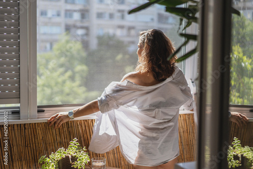 Young smiling caucasian woman in a white shirt standing on the balcony in the morning meeting a new day. View of summer city