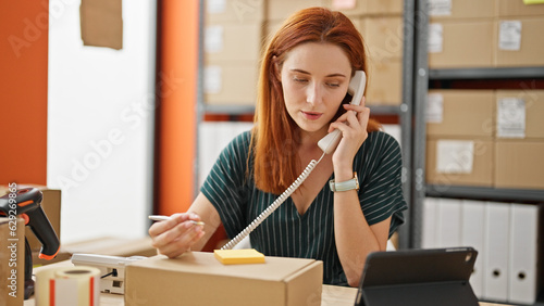 Young redhead woman ecommerce business worker talking on telephone writing on reminder paper at office