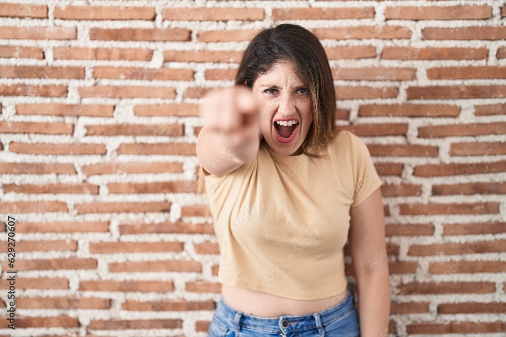 Young brunette woman standing over bricks wall pointing displeased and frustrated to the camera, angry and furious with you Young brunette woman standing over bricks wall pointing displeased and frustrated to the camera, angry and furious with you
