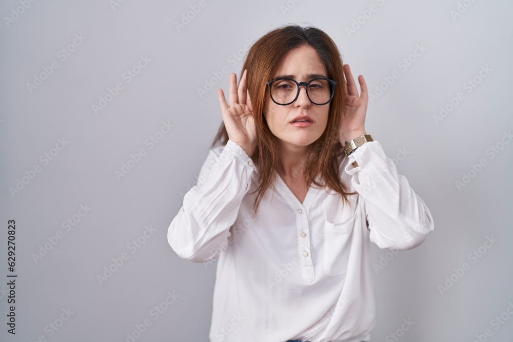 Brunette woman standing over white isolated background trying to hear both hands on ear gesture, curious for gossip. hearing problem, deaf Brunette woman standing over white isolated background trying to hear both hands on ear gesture, curious for gossip. hearing problem, deaf