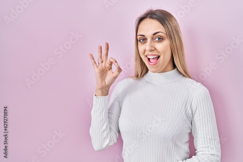 Young blonde woman wearing white sweater over pink background smiling positive doing ok sign with hand and fingers. successful expression.