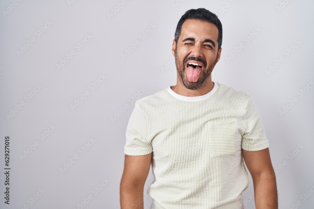Hispanic man with beard standing over isolated background sticking tongue out happy with funny expression. emotion concept. Hispanic man with beard standing over isolated background sticking tongue out happy with funny expression. emotion concept.