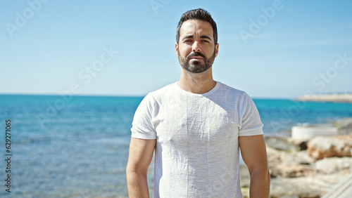 Young hispanic man standing with serious face at the beach