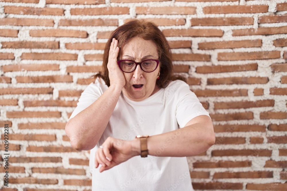 Senior woman with glasses standing over bricks wall looking at the watch time worried, afraid of getting late Senior woman with glasses standing over bricks wall looking at the watch time worried, afraid of getting late