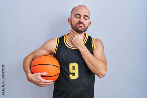 Young bald man with beard wearing basketball uniform holding ball touching painful neck, sore throat for flu, clod and infection