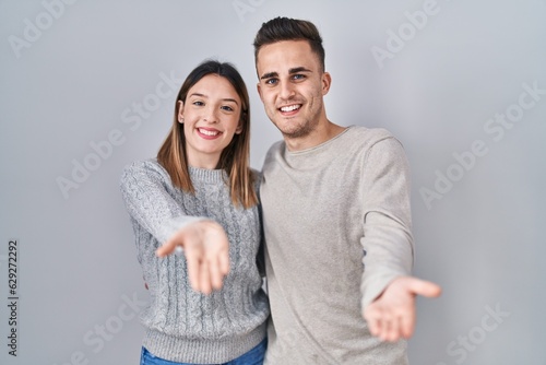 Young hispanic couple standing over white background smiling cheerful offering palm hand giving assistance and acceptance. © Krakenimages.com