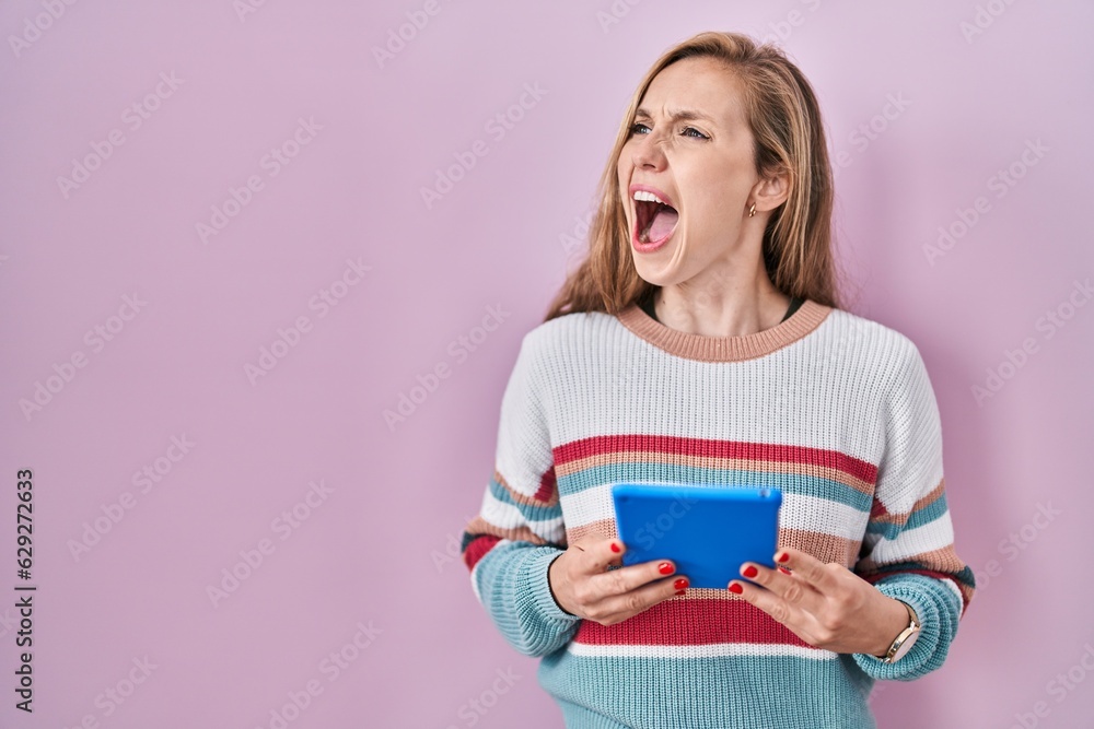 Young blonde woman holding touchpad angry and mad screaming frustrated and furious, shouting with anger. rage and aggressive concept. Young blonde woman holding touchpad angry and mad screaming frustrated and furious, shouting with anger. rage and aggressive concept.