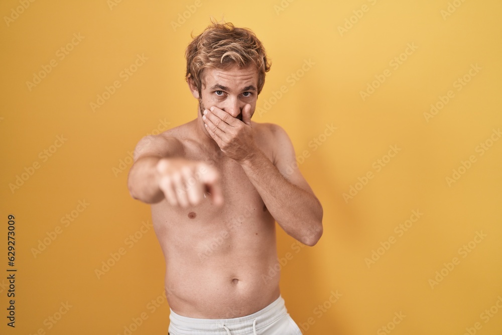 Caucasian man standing shirtless wearing sun screen laughing at you, pointing finger to the camera with hand over mouth, shame expression