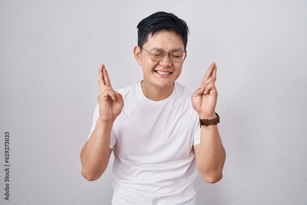 Young asian man standing over white background gesturing finger crossed smiling with hope and eyes closed. luck and superstitious concept. Young asian man standing over white background gesturing finger crossed smiling with hope and eyes closed. luck and superstitious concept.