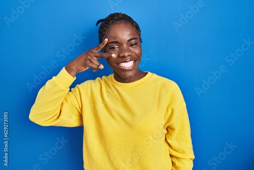 Beautiful black woman standing over blue background doing peace symbol with fingers over face, smiling cheerful showing victory