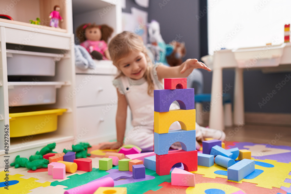 Fototapeta premium Adorable blonde girl playing with construction blocks sitting on floor at kindergarten