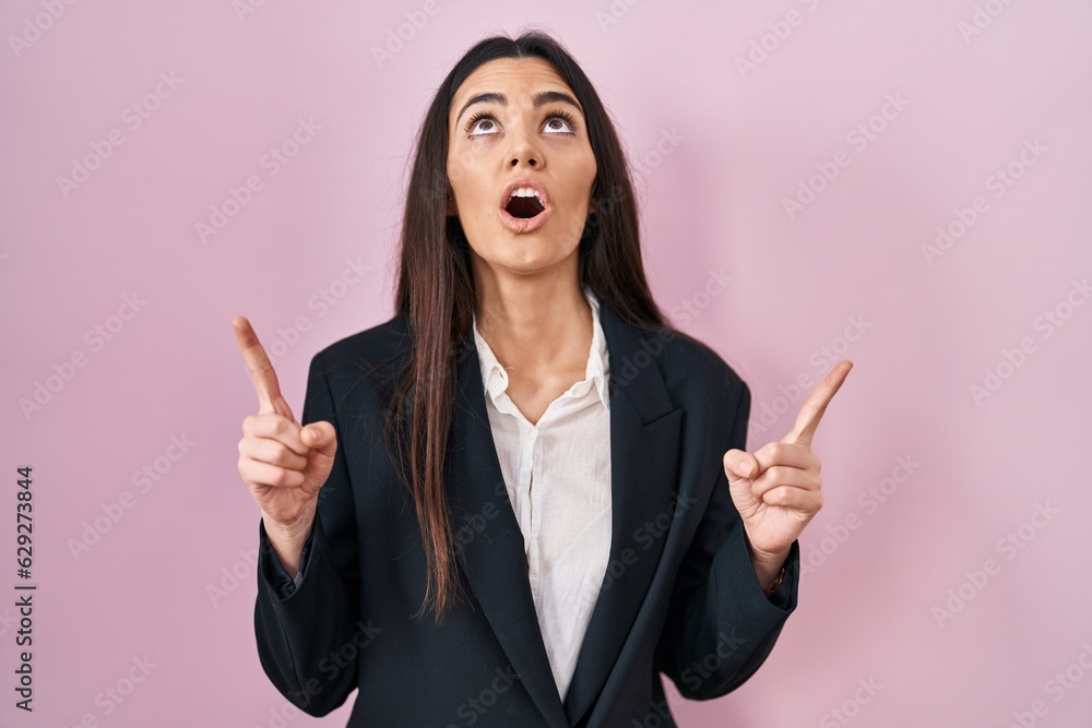 Young brunette woman wearing business style over pink background amazed and surprised looking up and pointing with fingers and raised arms. Young brunette woman wearing business style over pink background amazed and surprised looking up and pointing with fingers and raised arms.