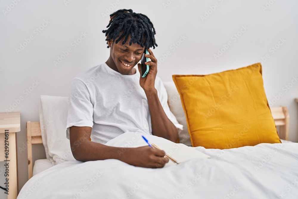 African american man talking on the smartphone writing on notebook sitting on bed at bedroom African american man talking on the smartphone writing on notebook sitting on bed at bedroom
