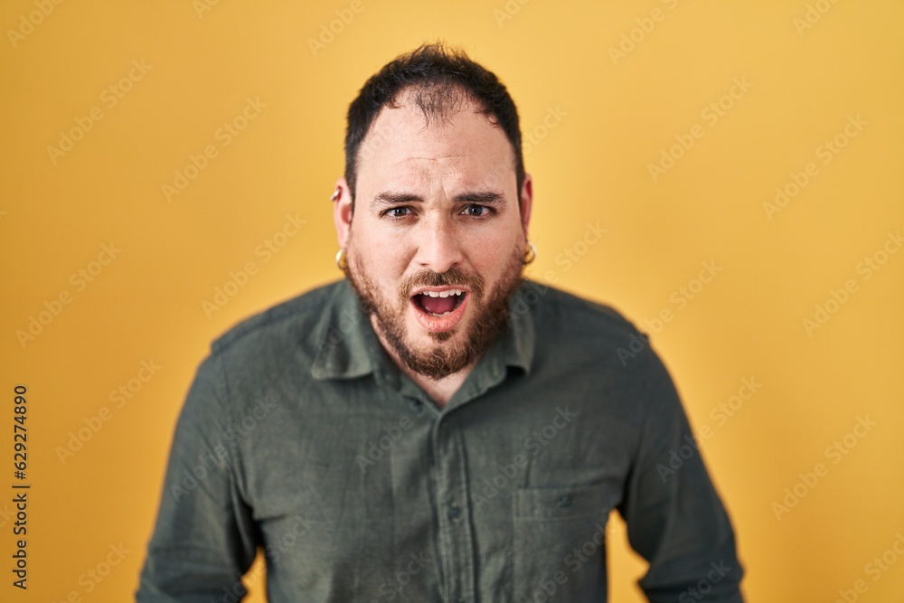 Plus size hispanic man with beard standing over yellow background afraid and shocked with surprise and amazed expression, fear and excited face. Plus size hispanic man with beard standing over yellow background afraid and shocked with surprise and amazed expression, fear and excited face.