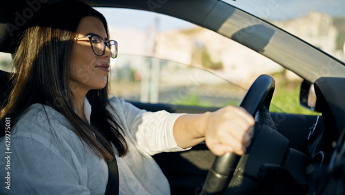 Young beautiful hispanic woman driving a car smiling wearing glasses on the road