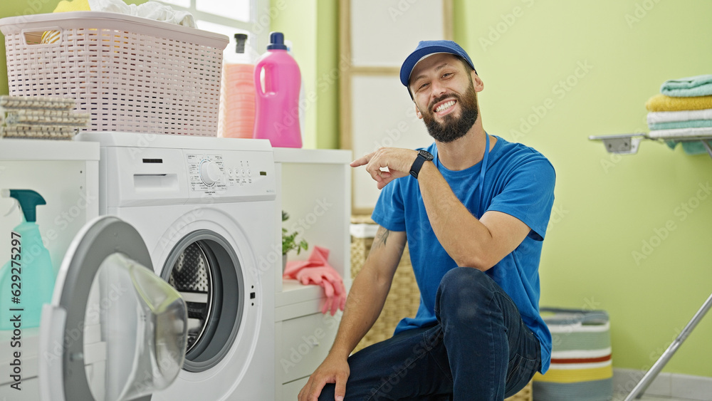 Young hispanic man technician pointing to washing machine smiling at laundry room