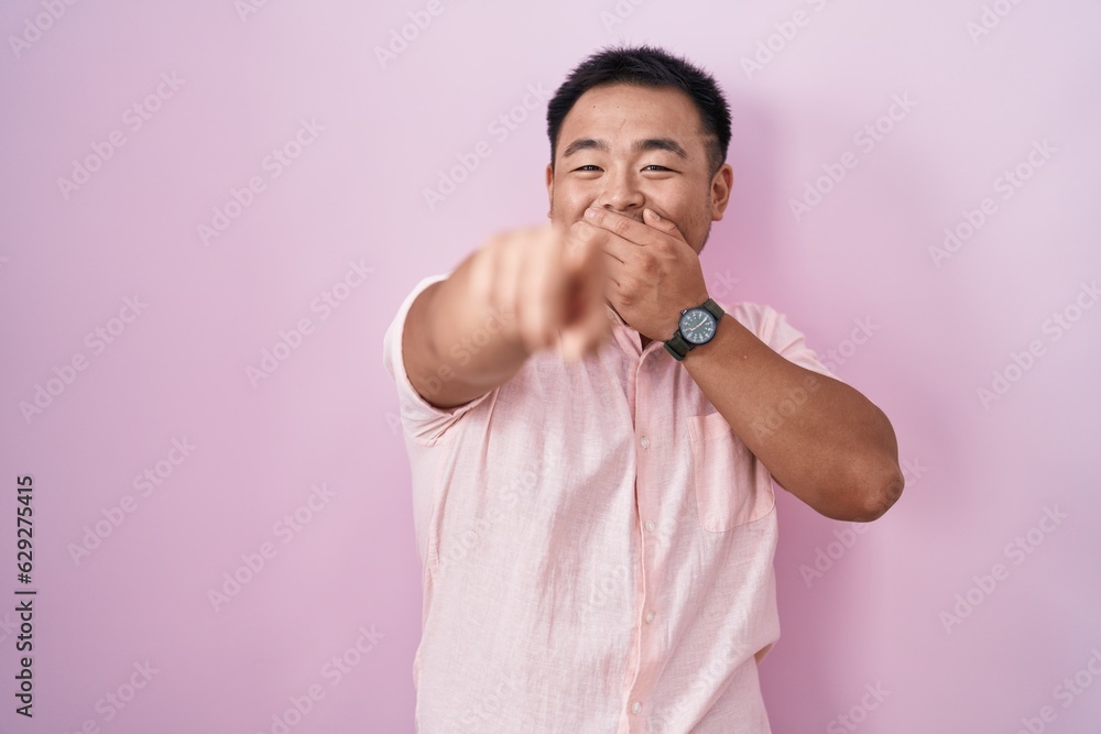 Chinese young man standing over pink background laughing at you, pointing finger to the camera with hand over mouth, shame expression