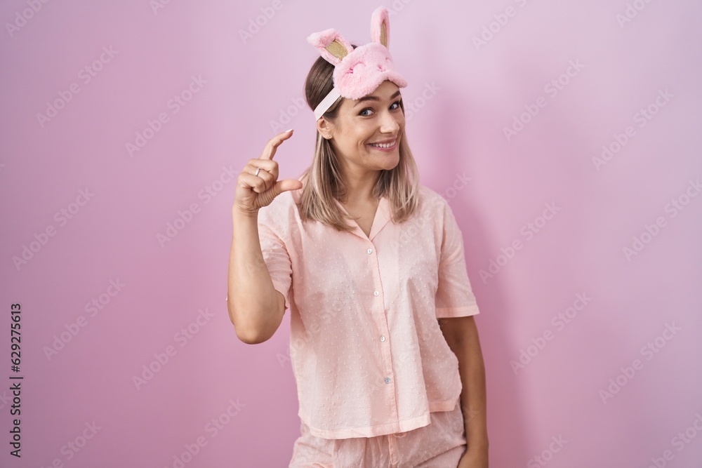 Blonde caucasian woman wearing sleep mask and pajama smiling and confident gesturing with hand doing small size sign with fingers looking and the camera. measure concept. Blonde caucasian woman wearing sleep mask and pajama smiling and confident gesturing with hand doing small size sign with fingers looking and the camera. measure concept.