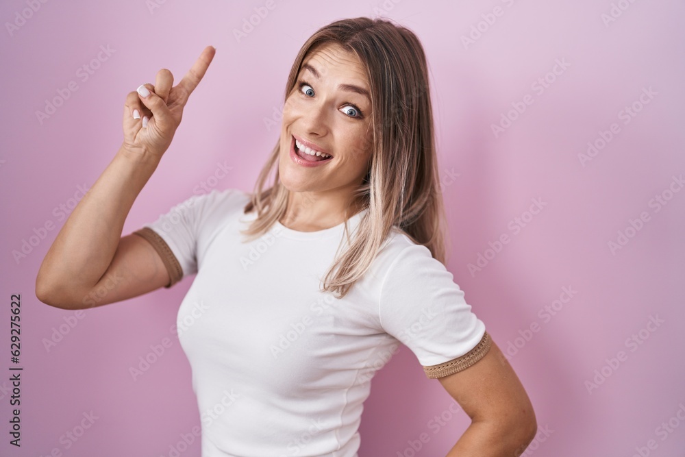 Blonde caucasian woman standing over pink background pointing finger up with successful idea. exited and happy. number one. Blonde caucasian woman standing over pink background pointing finger up with successful idea. exited and happy. number one.