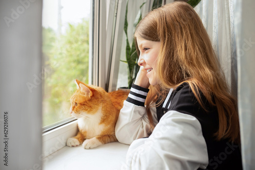 a cute girl laughs and looks out the window along with a ginger cat. friendship between child and pet