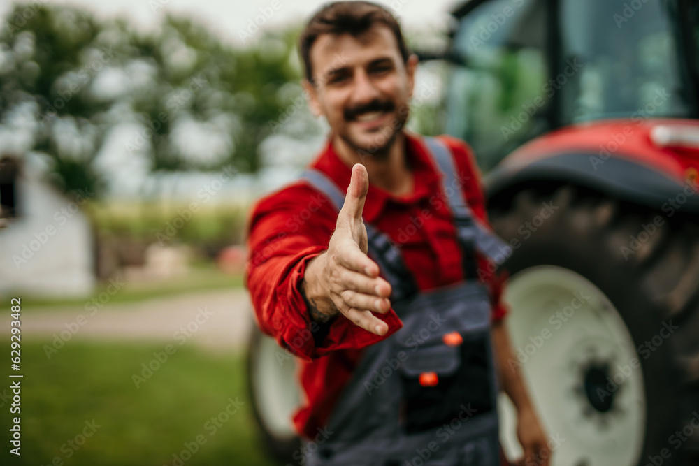 Smiling man in a working suit standing in front of a tractor and extending a hand for a handshake at the camera Smiling man in a working suit standing in front of a tractor and extending a hand for a handshake at the camera