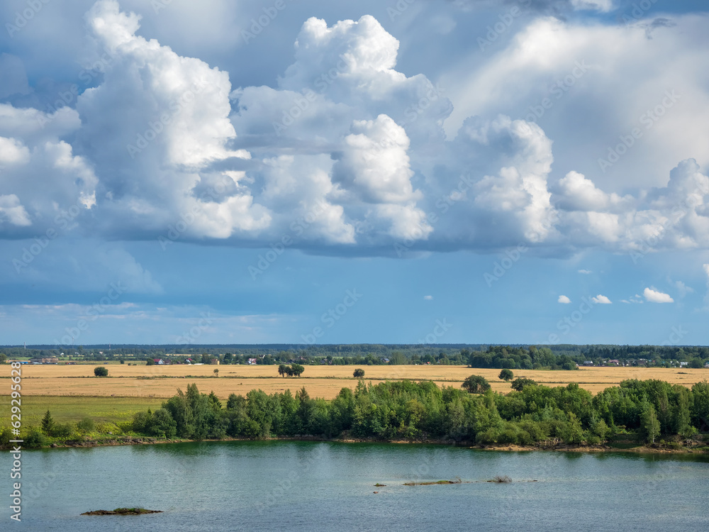 Amazing landscape with large white cumulus clouds over the lake and plain. Summer landscape on the banks of the green river at sunset.