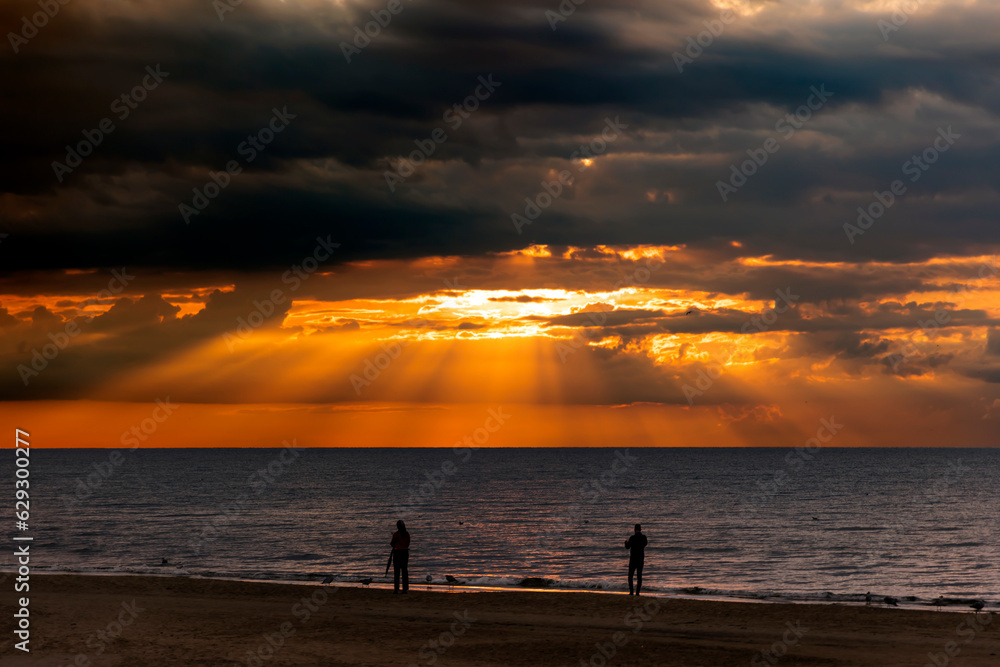 Silhouettes of people on the background of magic sunset with dark storm clouds and penetrating sunbeams above the Baltic sea in Jurmala, Latvia