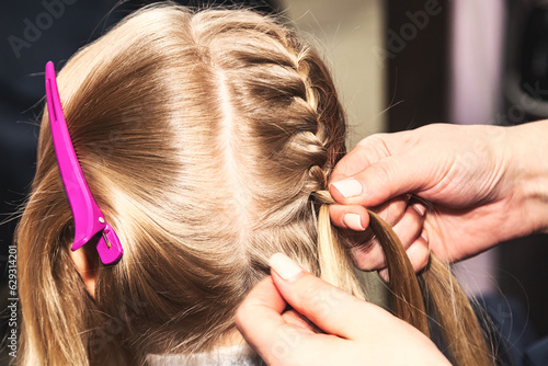 Hairdresser female hands makes braids for kid at barber shop, closeup. Hair salon, hair stylist braiding braid for lovely blonde little girl in barbershop. Haircare, hairdo concept. Copy ad text space