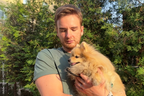 Young man holding on hands his Pomeranian Spitz dog, walking with cute puppy at summer day 
