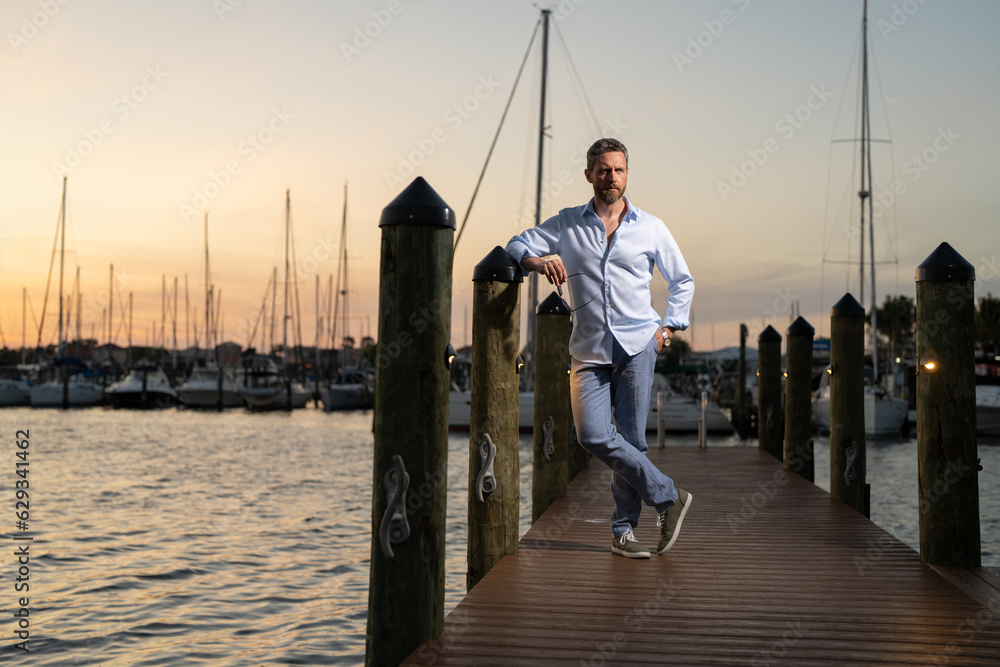 Millennial 40s man near the yacht. Portrait of fashion model outdoor. Fashion male posing near yachts. Fashion man in urban style. Handsome hispanic man at yacht club. Man in gulf near luxury yacht.