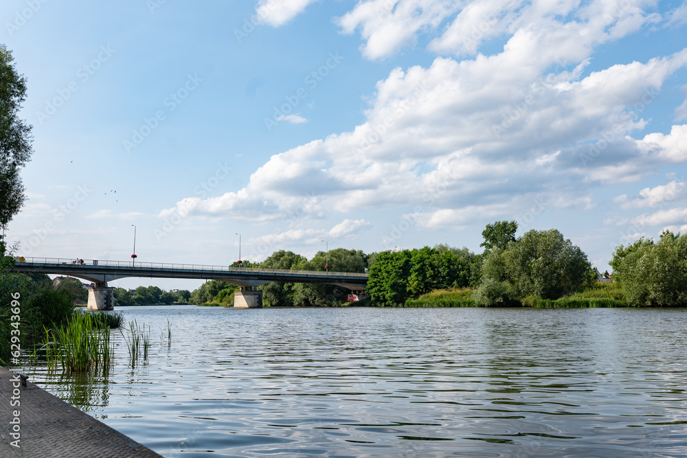 Rzeka Odra, w tle most lekko pochmurna pogoda, błękit nieba zieleń przy brzegu, pora letnia, Odra w odcinku województwa Opolskiego
