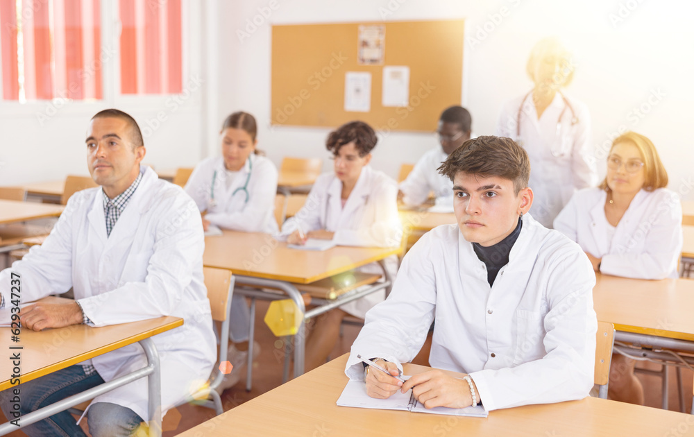 Group of interested young adult doctor trainees in white coats sitting at desks in university classroom, listening to lecture on general medical practice during refresher course..