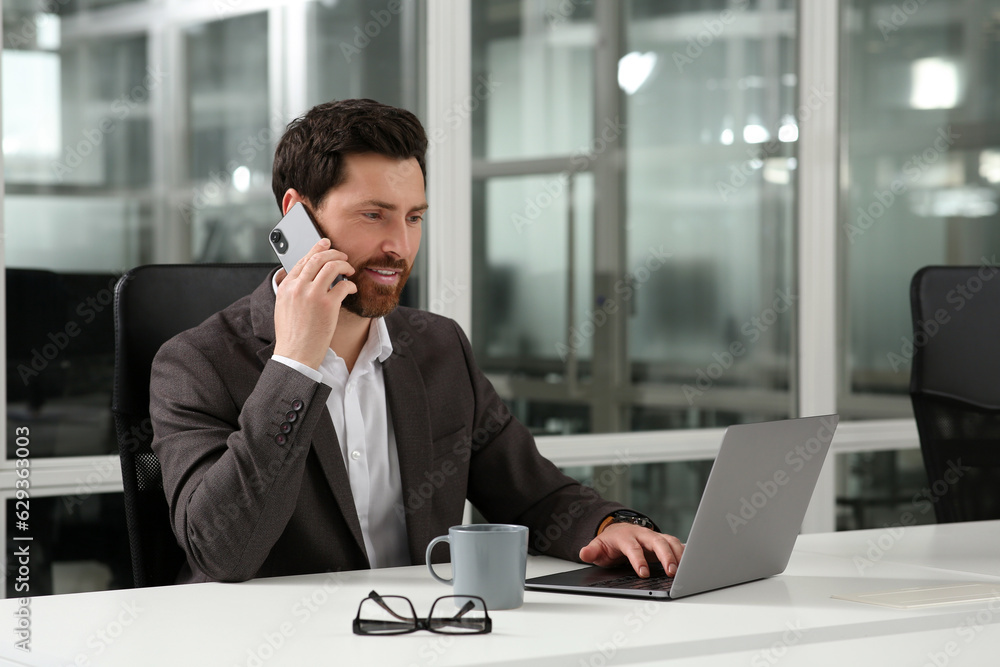 Man talking on phone while working with laptop at white desk in office Man talking on phone while working with laptop at white desk in office