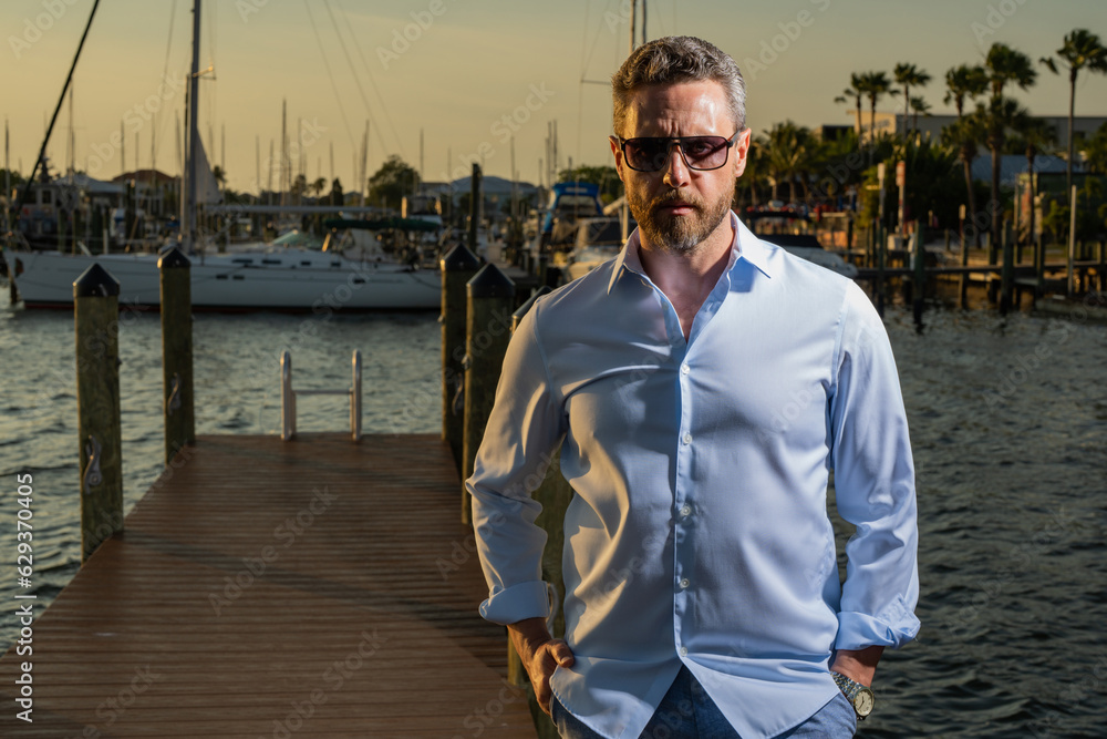 Rich stylish business man dreaming and thinking near the yacht. Portrait of confident man in yacht club. Stylish lambersexual model. Sexy man dressed in shirt. Fashion male posing on marina near yacht Rich stylish business man dreaming and thinking near the yacht. Portrait of confident man in yacht club. Stylish lambersexual model. Sexy man dressed in shirt. Fashion male posing on marina near yacht